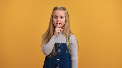Portrait smiling mystery cute little girl showing shh quiet gesture with finger near mouth posing isolated on orange studio. Adorable female kid shhh silence secret face expression forefinger at lips
