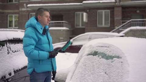 Person driver cleans the car from snow with a brush. A car covered in white snow after a winter snowfall or snow storm. Frozen vehicles in the parking lot.