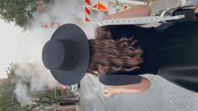 Vertical video. A young woman in a black dress and hat walks along the city street. Road works, orange barriers. Smoke or steam from the chimney. New York, USA. Accident, breakage of a heating pipe.