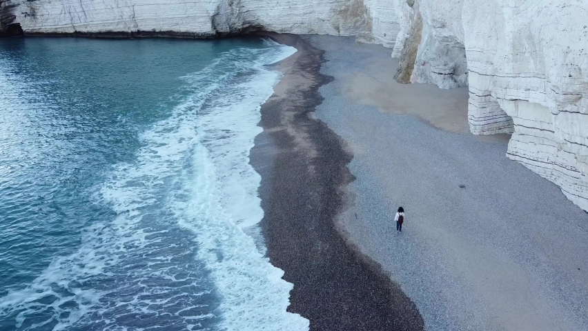 A girl walks alone on the beach and the drone chases her. The girl is so small compared to the white slopes of the cliff and the immensity of the sea waves | Shutterstock HD Video #1085583269