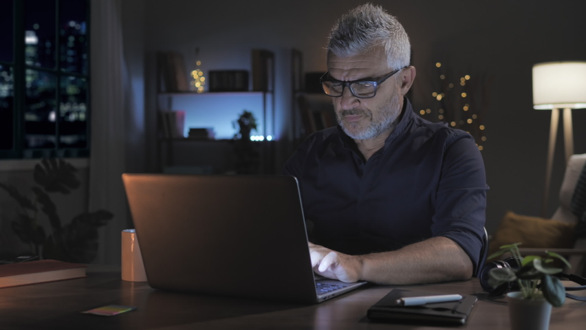 overwhelmed stressed mature man working late at night at the computer from home,caucasian middle aged male at the desk works at the laptop feels tired takes off eyeglasses rubs his eye Royalty-Free Stock Footage #1085589194
