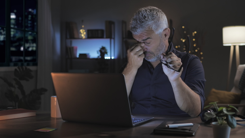 Overwhelmed stressed mature man working late at night at the computer from home,caucasian middle aged male at the desk works at the laptop feels tired takes off eyeglasses rubs his eye | Shutterstock HD Video #1085589194