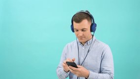Studio portrait of man in a blue shirt chooses music and turns it on in headphones from a smartphone. Cheerful office worker dancing in front of a bright blue background