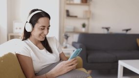 Happy positive young adult woman using a mobile phone while listening to music with headphones sitting on sofa at home. High quality 4k footage