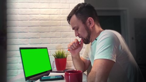 Man in front of a brick wall with a beard and in a white T-shirt works in the office or at home. Tired of work and watching the screen. Business and money, bitcoin, exchange. Laptop with green screen.