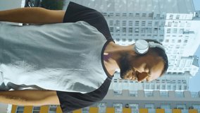 Bearded young man smiling on camera while standing on the balcony, and crossing arms. Vertical portrait of positive male person. Caucasian guy in wireless headphones outdoor, urban city
