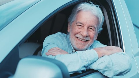 Happy owner looking at the camera with happy face. Handsome bearded mature man sitting relaxed in his newly bought car looking out the window smiling joyfully. One old senior driving and having fun.