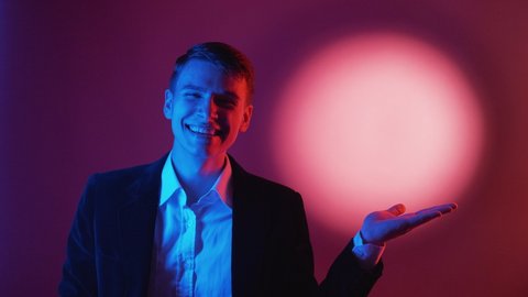 Advertising product. Happy man. Holiday surprise. Neon light male portrait in black suit holding opened palm posing red blue halo spotlight background.