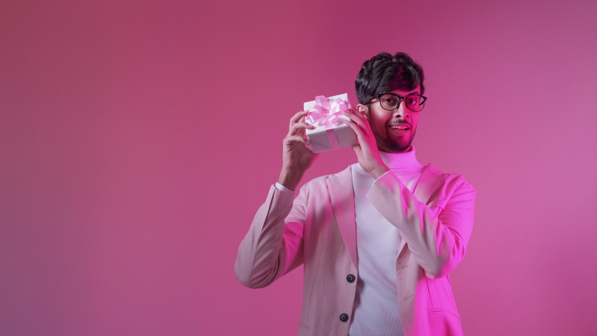Valentine day gift. Holiday surprise. Romantic present. Neon light curious joyful smiling handsome man in glasses showing box isolated on pink color copy space background. Royalty-Free Stock Footage #1085595788