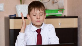 Theme of the online lesson on distance learning for schoolchildren. Caucasian boy 7-8 years old at home raises his hand to answer the teacher's question, holds a gadget at an online conference lesson.