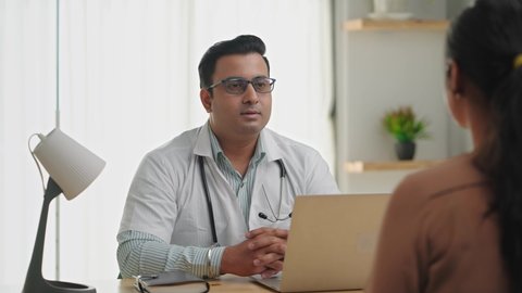 An Asian Indian male smiling physician or doctor sitting in a modern clinic wearing a stethoscope and apron giving consultation to a patient. Medical, medicine, and healthcare concept