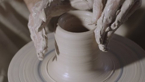 The potter's male hands shape and sculpt the soft white clay pot, spinning on a potter's wheel in a creative workshop. The master gives a shape to the pottery. Clay shaping close up. Slow motion.
