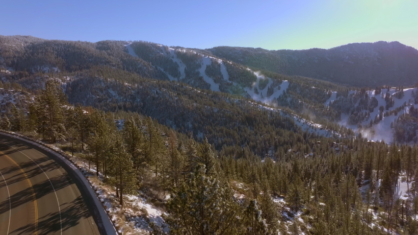 Boom down towards road winding through mountains in Lake Tahoe, Nevada on a beautiful day with Douglas Firs in the foreground and mountain peaks on the horizon.