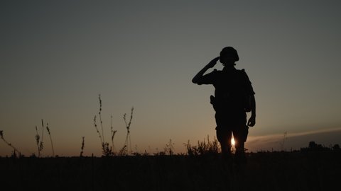 Silhouette of fully equipped soldier in combat helmet and ammunition saluting during sunset on field, special forces fighter stands on background of colorful sky and shows respect by greeting officer.