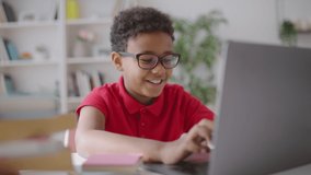 Cheerful schoolboy doing online hometask, typing on laptop at home, education