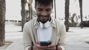 Front view of young man using cellphone outdoors - Cheerful hispanic guy chatting on mobile phone while sitting on bench - Social media, communication and millennial generation people concept. 