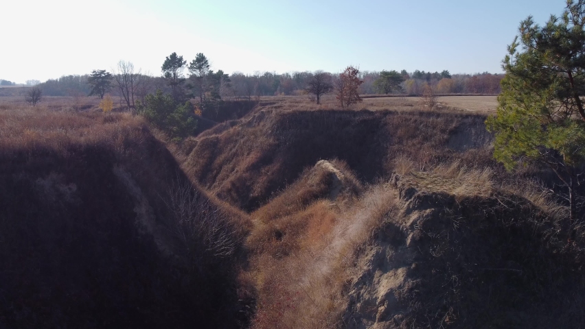 Deep ravine with steep clay slopes, overgrown with dry grass and single trees, aerial view while moving forward
