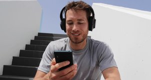 Podcast. Man listening to podcasts or audiobook on mobile phone app wearing headphones sitting outdoors on stairs. Healthy lifestyle sport athlete using smartphone on break outsite relaxing