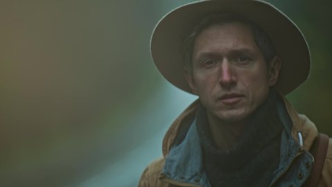 close up portrait of man hitchhiker wearing in retro style. traveler in hat with backpack stands outdoors at road in cold wet foggy autumn day and waites a car. soft focus