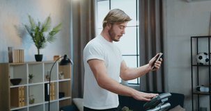 Likable active sportive young bearded man in white t-shirt jogging on treadmill at home and talking with friend via phone video chat