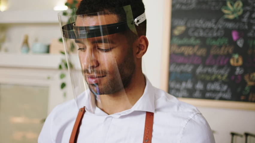 Cozy coffee shop closeup to the camera portrait of a barista with protective mask looking to the camera smiling cute Royalty-Free Stock Footage #1085625278