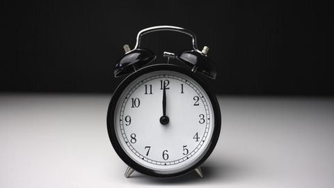 A black vintage bell quartz analog alarm clock in a black background is ringing at 12 noon midnight.