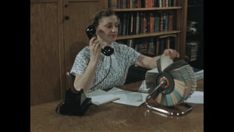 1950s: Woman answers phone at desk in AV Library office. Men and women enter office and talk.