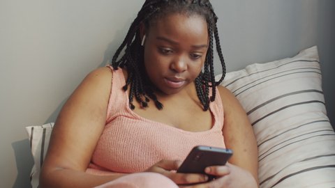 Horizontal medium shot of attractive young Black woman sitting relaxed on bed in morning listening to music in earphones and surfing Internet on smartphone