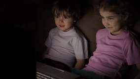 Boy and Girl Watch Cartoon on Laptop on Living Room. Concept Video Game, Entertainment, Emotions, Family. Children Brother and Sister Watching TV. Portrait Сute Little Kids While Watching TV on Laptop