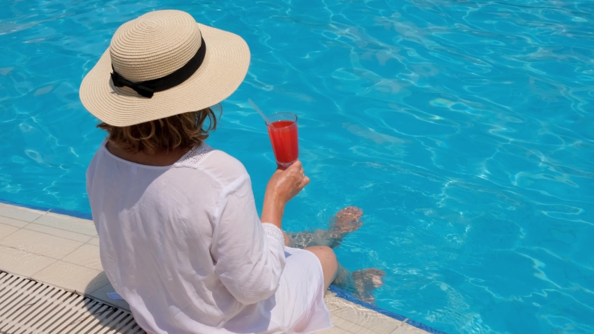 Woman in a straw hat relaxes near the swimming pool, drinks a pink cocktail and dangles her legs in the water. Summer mood concept | Shutterstock HD Video #1085636447