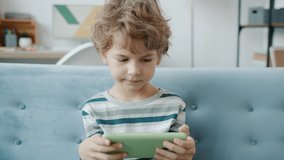 Slow motion of joyful kid having fun with video game on smartphone playing alone sitting on couch at home. Modern devices and indoor entertainment concept.
