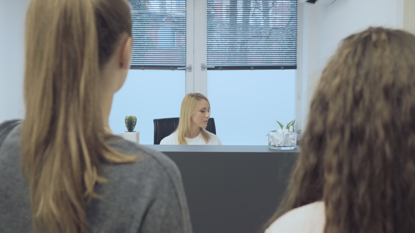 Over the shoulder of two female patients standing at the reception desk in a medical office and talking with a Caucasian female receptionist. Royalty-Free Stock Footage #1085639312