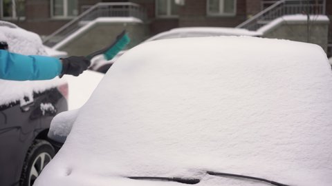Person driver cleans the car from snow with a brush. A car covered in white snow after a winter snowfall. Frozen vehicles in the parking lot.