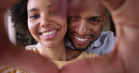 Affectionate African American couple in love enjoy romance in life look at web camera through heart shape of united palms. Romantic spouses shoot self video record greeting with Valentine Day. Closeup