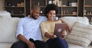 Relaxed millennial African American couple cuddle on sofa hold digital touchpad device choose consumer goods in convenient ecommerce app. Happy young spouses enjoy leisure time at home with tablet pc