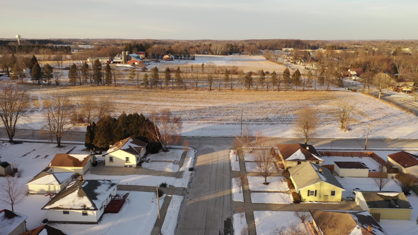 Aerial above houses covered in snow in a small town in USA in winter, farm and agricultural fields in  background. Suburban neighborhood in Midwest USA. Establishing shot of America