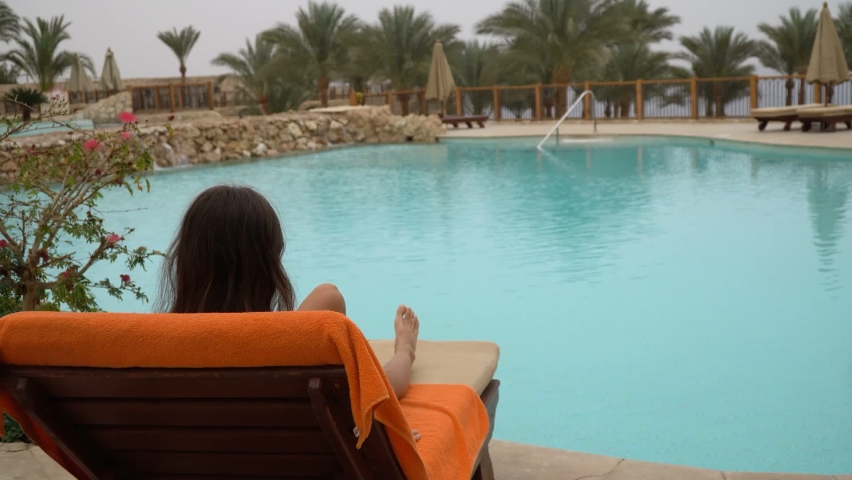 Young beautiful woman sitting on sun loungers by swimming pool at the hotel | Shutterstock HD Video #1085647424