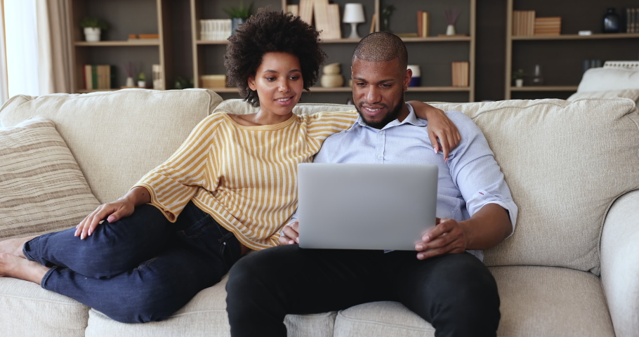 Happy Black couple rest on cozy couch hold laptop plan home renovation choose interior design online discuss buying furniture at web store. Young husband wife browse websites on computer talk have fun Royalty-Free Stock Footage #1085648378