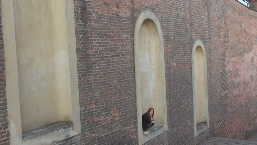 She is sitting in a stone place. Goodbye waiting. Venue. Brick wall. A woman is waiting for a meeting with a friend in the old city.