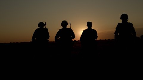 Group of soldiers in combat helmets and ammunition walk across field, silhouettes of special operations forces infantrymen returning to homeland on sunset, shooters protect country from hostile fire.