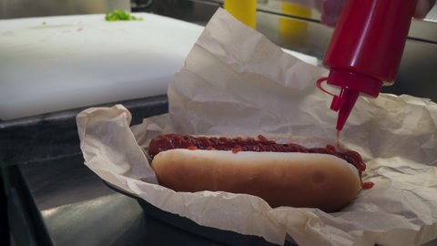 Preparing Hot Dog By Spreading Red Ketchup Sauce On Hot Grilled Sausage. Cook Pouring Ketchup Sauce From Bottle On Hot Dog. Adding Ketchup To Tasty Hot Dog. Fast Food. American Cuisine Dish