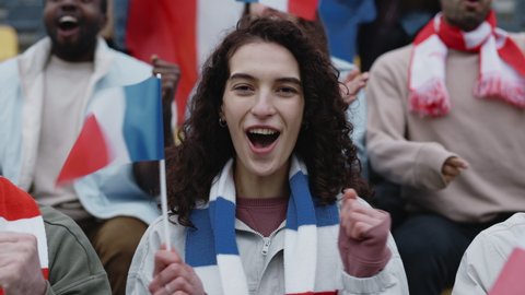 Portrait of pretty woman with dark curly hair gesturing and screaming from bleachers while watching soccer game. French flag in hands and scarf on neck.