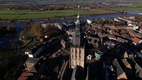 Slow aerial rotation around Walburgiskerk church tower in picturesque town of Zutphen in The Netherlands with a ship vessel passing on the river IJssel in the background contrasted with floodplains