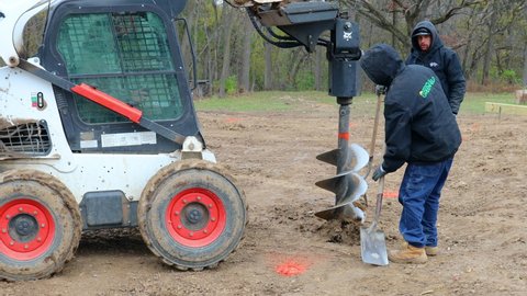 Cambridge , Illinois , United States - 11 16 2021: Three construction workers working with shovels and hydraulic auger mounted on skid steer loader to dig a post hole