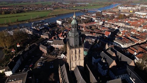 Slow aerial rotation around Walburgiskerk church tower in picturesque town of Zutphen in The Netherlands following a cargo ship vessel passing by on the river IJssel in the background