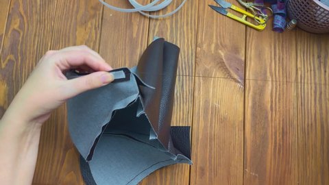 Woman sews plastic ribbon with thread and needle to clothing made of eco-leather. Workshop of seamstress. Neck corset for cosplay or costume. Scissors on wooden table.