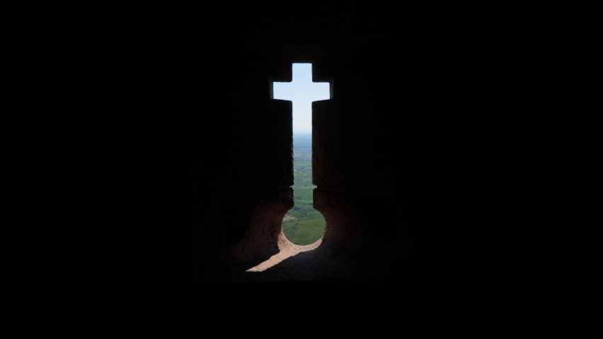 Crosslet loop or embrasure in wall of old castle, green landscape seen through narrow opening. Firing slot in shape of inverted keyhole with cross, old fortification on top of hill