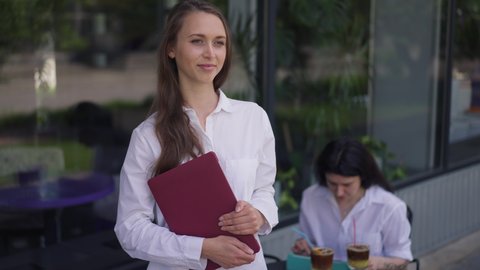Portrait of cute confident young woman posing outdoors in sidewalk cafe with blurred friend sitting at table at background. Happy smiling Caucasian millennial lady with laptop looking at camera