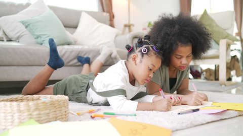 Happy African family cute child girl kid sibling lying on the floor using color pencil drawing and painting on paper book together. Little sister enjoy and fun indoor leisure activity together at home