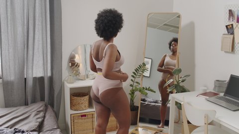Rear-view tracking slowmo shot of curvy African-American woman in underwear standing in front of mirror in bedroom measuring her waist with tape measure